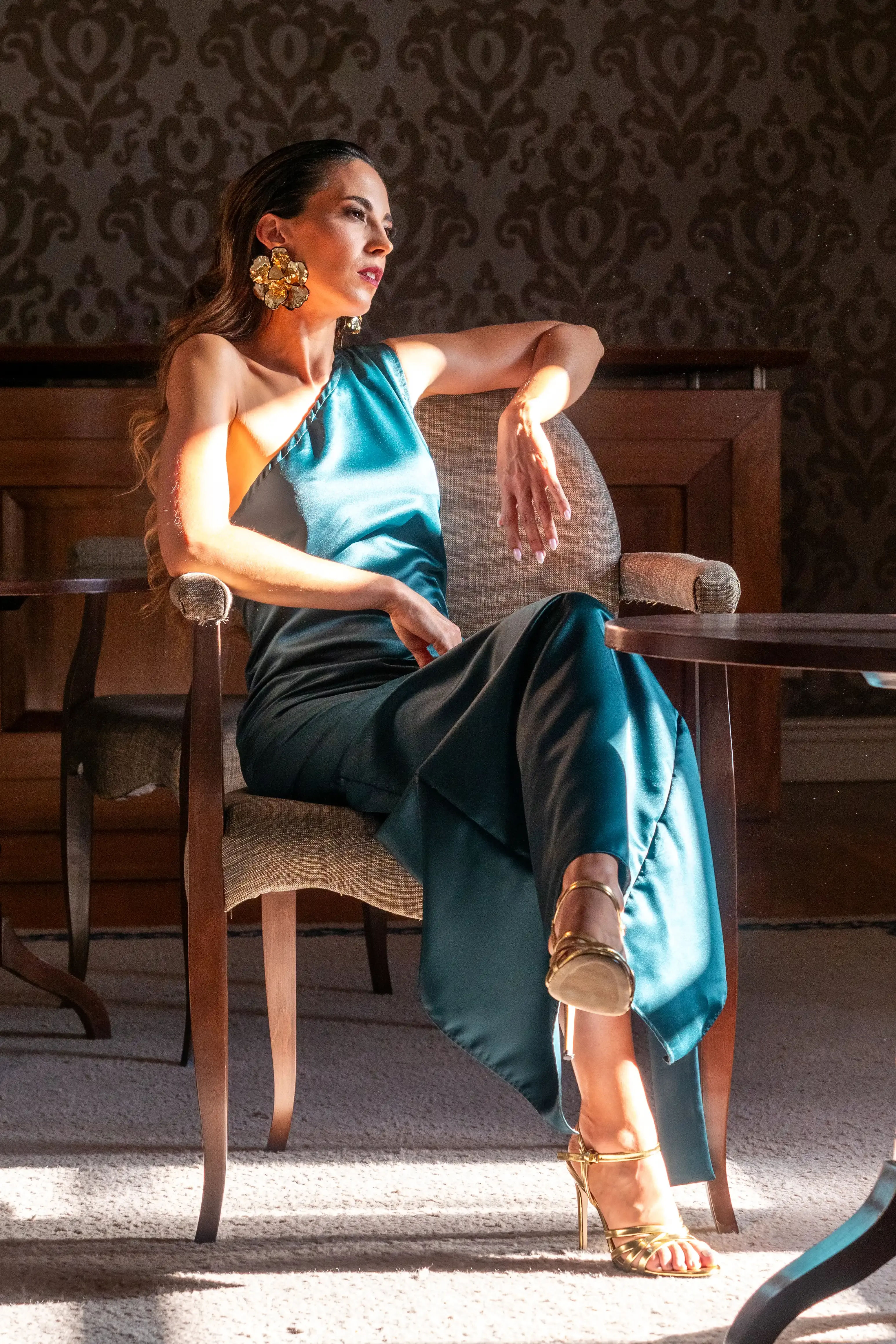 Uma mulher posa com um vestido comprido e azul sentada na cadeira iluminada pela luz solar direta.